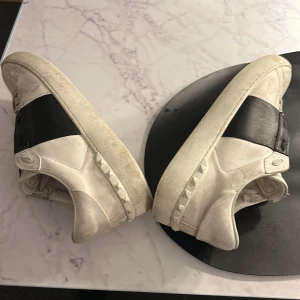 Valentino opens  - Säljer ett par vita  sneakers med ett brett svart band över snörningen. Skorna har en cool och stilren design med nitar runt sulan. De är i bra skick men har några märken från användning. Perfekta för en casual look eller att dressa upp en outfit.