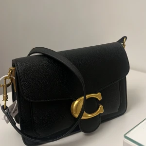 Svart coach väska med guld detaljer , helt ny -  svart coach axelväska i skinn med en stor guldfärgad spänne framtill. Perfekt för både vardag och fest. Väskan har en justerbar axelrem och är i nytt skick. En stilren och tidlös design. Pris kan diskuteras 