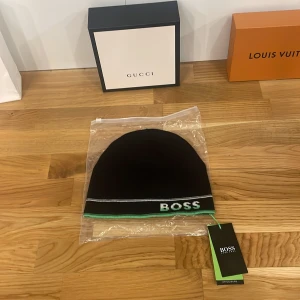 Svart mössa från BOSS - Snygg svart mössa från BOSS med gröna och vita detaljer. Perfekt för att hålla sig varm och stilren under kyliga dagar. Mössan är i nytt skick och kommer i originalförpackning. Passar perfekt för både vardag och mer uppklädda tillfällen.