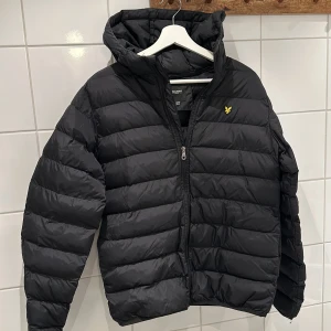 Svart pufferjacka från Lyle & Scott - Säljer en svart pufferjacka från Lyle & Scott i storlek 15/16 år. Jackan har en mysig huva och praktisk dragkedja framtill. Perfekt för kyliga dagar och håller dig varm och bekväm. Den är i mycket bra skick och redo för nya äventyr! 🖤