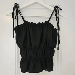 Svart ärmlös topp med knytband - Snygg svart topp med knytband på axlarna. Den är ärmlös och har en elastisk midja & passar därav stl XS-M. Perfekt för en kväll ute! Aldrig använd - nyskick