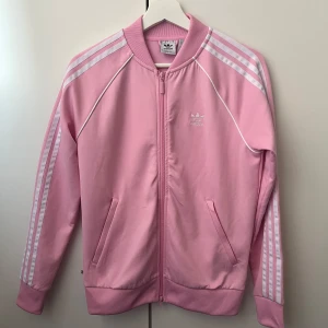 Rosa jacka från Adidas - Säljer en snygg rosa jacka från Adidas i storlek S. Den har de klassiska tre ränderna på ärmarna och en dragkedja framtill. Perfekt för vår och sommar! Jackan är i mycket bra skick och gjord av återvunnet material.