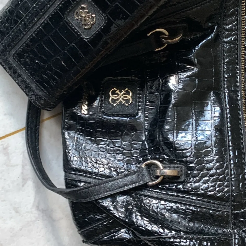 Snygg svart axelväska och matchande plånbok från Guess i krokodilmönstrat skinn. Väskan har en praktisk dragkedja och stilrena detaljer i silver. Perfekt för både vardag och fest!. Laukut & Käsilaukut.