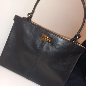Svart skinnväska från Wera - Snygg svart skinnväska från Wera i mycket bra skick. Den har en elegant design med guldfärgad låsdetalj och bekväma handtag. Perfekt för både vardag och fest. Kommer med en skyddande dustbag. Har aldrig använt för den var inte riktigt min stil men den är ungefär max en vecka gammal men råkade slänga kvittot, kostade 1299kr ny.