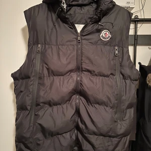 Svart dunväst från Moncler - Säljer en svart dunväst från Moncler. Västen har en dragkedja och två fickor med dragkedjor. Den har hål vid under armen som lätt går att sy ihop samt ena fickan. Även vid skulderbladet. Perfekt för höst och vinter, håller dig varm och stilren. Passar bra till både vardag och fest.