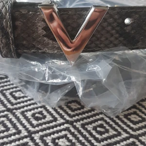Svart bälte från Valentino - Säljer ett snyggt svart bälte från Valentino i skinn med en cool V-formad spänne i silver. Bältet har en textur som påminner om ormskinn vilket ger det en lyxig känsla. Perfekt för att piffa upp vilken outfit som helst! 🖤