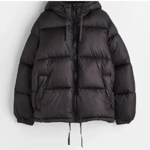 Svart pufferjacka med huva - Säljer en svart pufferjacka i syntetmaterial med huva och dragkedja. Jackan är perfekt för kalla dagar och har en oversized passform som ger en cool look. Den är långärmad och har praktiska fickor på sidorna. Perfekt för både höst och vinter!