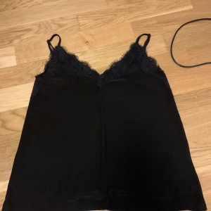 Svart linne med spetsdetaljer - Säljer ett snyggt svart linne med spetsdetaljer vid axelbanden och halsringningen. Perfekt för både vardag och fest. Linnet är i bra skick och har en bekväm passform.