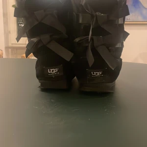 Svarta boots med rosetter från UGG - Säljer ett par svarta UGG boots i bra skick. De har en mysig pälsfodring och fina rosetter på baksidan. De är i storlek 38/39