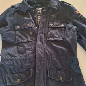 Field jacket  - Säljer en snygg mörkblå field jacket från NZA New Zealand Auckland. Jackan har flera fickor med knappar och en dragkedja framtill. Den har också en cool broderad detalj med flagga på ärmen. Perfekt för höst och vår! Pris ej hugget i sten.