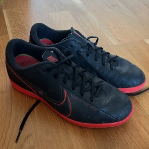 Svarta Nike fotbollsskor - Säljer ett par svarta Nike fotbollsskor i bra skick. De har en snygg röd sula och Nike-logga på sidan. Skorna har snörning och är perfekta för inomhusfotboll. Passar dig med storlek EUR 38. Skorna är bekväma och ger bra grepp på planen.