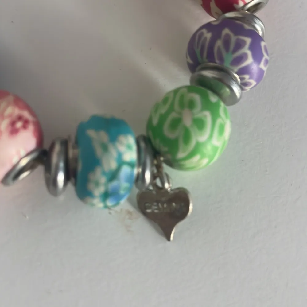 Säljer ett superfint armband med stora, färgglada pärlor i olika färger som röd, blå, grön och lila. Varje pärla har ett blommigt mönster och är separerade av silvriga mellandelar. Perfekt för att liva upp vilken outfit som helst! 💐. Asusteet.