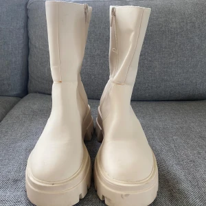 Beige boots med dragkedja - Säljer ett par snygga beige boots i syntetmaterial. De har en chunky sula och dragkedja på sidan för enkel på- och avtagning. Perfekta för höst och vår, dessa boots ger en cool look till vilken outfit som helst. De är i mycket bra skick och redo för nya äventyr!