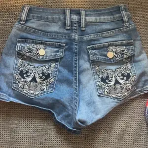 Snygga jeansshorts i blått med hög midja och coola broderade detaljer på bakfickorna. De har en knapp och dragkedja framtill och är i bra skick. Passar perfekt till en avslappnad stil. Köpt från SHEIN men används inte