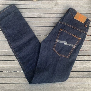 Mörkblå Nudie Jeans - Säljer ett par näst intill nya mörkblå nudie jeans i mycket bra skick. 28-32, skriv privat för mått. De har kontrastsömmar och en klassisk femficksdesign. Perfekta för både vardag och fest! Passformen är slim och de är midwaist. Sköna och stilrena, ett måste i garderoben!