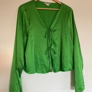 Grön satinblus med knyt - Säljer en superfin grön blus i satinmaterial. Den har långa ärmar och en v-ringad design med knyt framtill. Perfekt för både vardag och fest!