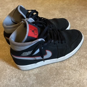 Svarta Nike Air Jordans - Snygga svarta Nike Air Jordans med grå och röda detaljer. Skorna har en klassisk design med snörning och en grå sula. Har ett litet märke på vänstra skon (sista bilden) men det stns knappt. Endast skor ingår.