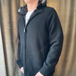 Fodrad Cardigan med päls - Säljer en svart stickad jacka med en mysig huva kantad med pälsluva. Jackan har dragkedja framtill och ribbade muddar vid ärmslut och nederkant. Perfekt för kyliga dagar! Vid minsta fråga eller fundering hör av er🤝