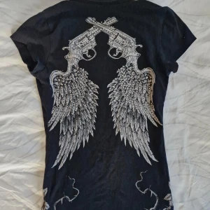 Alternativ t-shirt med rhinestones - Cool T-shirt från Rock & Rose! 🖤 V-ringad modell med rosor på framsidan, pistoler och vingar på ryggen. Även små rhinestones på baksidan.  Sann till storlek (S) I fint använt skick! :)
