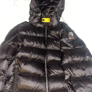 Svart dunjacka från Parajumpers - Säljer en snygg svart dunjacka från Parajumpers i modellen Sheen. Jackan har en hög krage och en praktisk huva. Den är quiltad med dragkedja framtill och har en ikonisk gul detalj vid halsen. Perfekt för kyliga dagar!