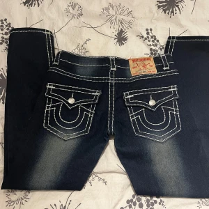 True Religion  - Snygga mörkblå jeans med vita kontrastsömmar och unika bakfickor med lock och knappar. Jeansen har en klassisk femficksdesign och en medelhög midja. Perfekta för en avslappnad stil.