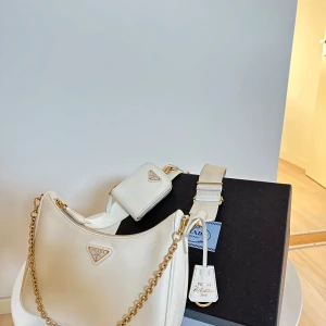 Vit axelväska från Prada - Elegant vit axelväska från Prada med guldfärgade kedjedetaljer och matchande axelrem. Väskan har en stilren design med en liten tillhörande plånbok. Perfekt för att ge en lyxig touch till din outfit. Kvitto och kartong finns.