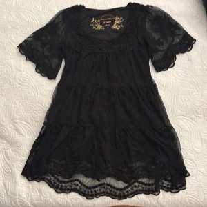 Svart klänning från KappAhl - Snygg svart klänning/blus från KappAhl med korta ärmar och spetsdetaljer. Tillverkad i polyamid med polyesterfoder, perfekt för en elegant look. Klänningen/blusen har en vacker struktur och en feminin siluett. Lik en klänning men väldigt kort, rekommenderar att bära kort byxor eller en kjol under 😊