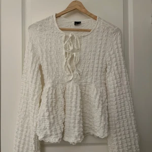 Vit långärmad blus med knyt - Charmig vit blus med lång ärm och knytning framtill. Den har en texturerad yta som ger en unik look. Perfekt för att lägga till en romantisk touch till din outfit.