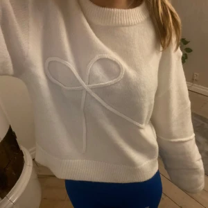 Vit stickad tröja med rosettdetalj - Mysig vit stickad tröja med en dekorativ rosett på framsidan. Tröjan har långa ärmar och ribbade kanter vid hals, ärmslut och nederkant. Supersnygg och prislappen sitter kvar!