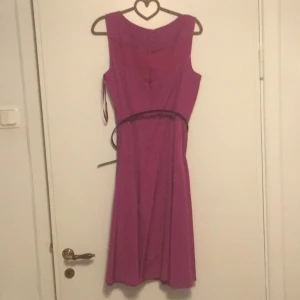 Fuchsia klänning från Monsoon - Snygg fuchsia klänning från Monsoon med en elegant design. Klänningen har en smickrande passform och ett smalt bälte i midjan. Perfekt för att lysa upp garderoben med en färgstark touch. Orginal pris 899kr