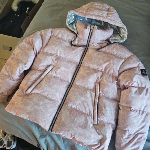 Rosa pufferjacka med huva - Säljer en rosa pufferjacka med huva och dragkedja. Jackan har ett subtilt mönster och är perfekt för kyligare dagar. Den har långa ärmar och praktiska fickor framtill. Äkta!!!! Priset kan diskuteras vid snabb affär