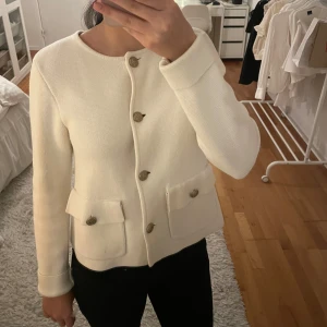 Vit kavaj/kofta - Elegant vit kavaj med rund hals och dekorativa knappar framtill. Den har två praktiska fickor och är tillverkad i ett strukturerat material som ger en stilren look. Perfekt för att lyfta vilken outfit som helst.
