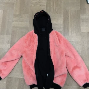 Rosa teddyjacka från Diadora - Säljer en snygg rosa teddyjacka från Diadora med svart huva och dragkedja. Jackan är gjord av polyester och akryl. Den har en cool och avslappnad stil med långa ärmar och en mysig känsla. Köpt i Italien och knappt använd