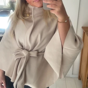 Kappa/poncho missäy  - Elegant beige kappa med knytband i midjan och dekorativa knappar. Den har en lös passform och vida ärmar, perfekt för en stilren look.