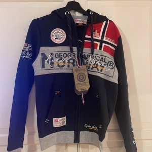 Navy blue hoodie från Geographical Norway - Säljer en snygg navy blue hoodie från Geographical Norway med dragkedja och tryck av norska flaggan. Tröjan har långa ärmar och flera broderade detaljer samt texttryck på ärmen och bröstet. Perfekt för lite kallare dagar! Nypris 1049kr, tröjan är bara provad. Hör gärna av er vid frågor eller funderingar😁
