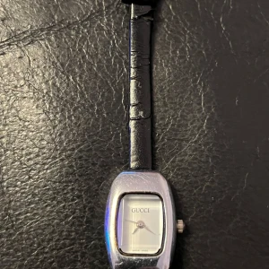 Vintage Elegant klocka från Gucci - Snygg Gucci-klocka med en stilren vit urtavla och silverfärgat hölje. Klockan har en svart skinnrem och är Swiss Made. Perfekt för den som vill ha en tidlös accessoar med lyxig känsla.