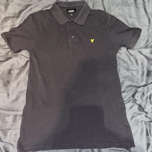 Grå pikétröja från Lyle & Scott - Säljer en stilren grå pikétröja från Lyle & Scott med korta ärmar och klassisk krage. Tröjan har en broderad gul logotyp på bröstet. Perfekt för en avslappnad och snygg look. Inte för varm inte för kall perfekt till en sommar dag i stan. Säljer då den är för liten