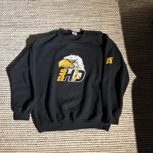 Svart sweatshirt från Gildan med örntryck - Säljer en svart sweatshirt från Gildan med ett stort tryck av en örn och texten '1340 HD' i gult och vitt på framsidan. Tröjan har långa ärmar och är tillverkad i en polyesterblandning för extra komfort.