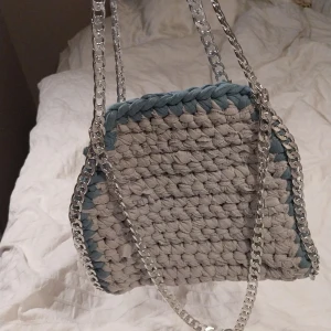 Grå virkad väska med kedjedetaljer - Snygg grå virkad väska med silverfärgade kedjedetaljer. Väskan har en unik design med blåa kanter som ger en fin kontrast. Perfekt för att ge din outfit en cool touch.