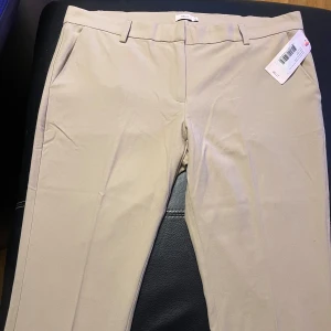 Beige kostymbyxor från Nelly - Snygga beige kostymbyxor från Nelly med en klassisk design. Byxorna har en rak passform och är perfekta för en stilren look. De har hällor för bälte och diskreta bakfickor.