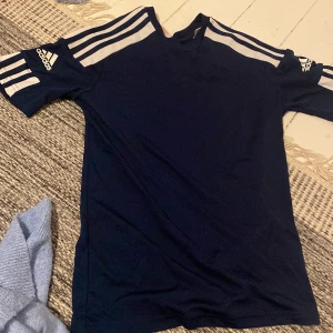 Mörkblå sporttröja från Adidas - Säljer en mörkblå sporttröja från Adidas med de klassiska tre ränderna i vitt på ärmarna. Tröjan har korta ärmar och en bekväm passform, perfekt för träning eller vardagsbruk.