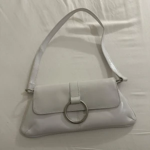 Vit axelväska med metallring - Snygg vit axelväska i syntetmaterial med en stor metallring som detalj på framsidan. Väskan har en enkel och stilren design med ett lock som stängs över öppningen. Perfekt för att bära det nödvändigaste med stil.