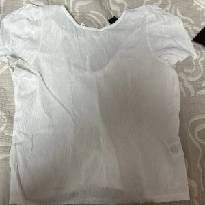 Vit ribbad topp från Lindex - Säljer en vit ribbad topp från Lindex med korta ärmar och knytning i ryggen. Perfekt för en stilren look.
