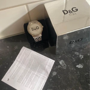 Silverfärgad klocka från D&G - Snygg och elegant klocka från D&G med en silverfärgad länk och glittrande detaljer runt urtavlan. Perfekt för att ge en touch av lyx till din stil. Levereras med originalförpackning och dokumentation. Skriv om du har några frågor 💕