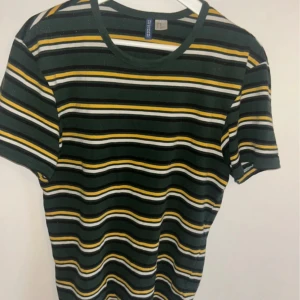 Randig t-shirt  - Säljer en cool randig t-shirt från H&M i grönt, svart, vitt och gult. Den har en klassisk rund hals och korta ärmar. Perfekt för en avslappnad stil!