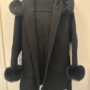Svart kappa med päls detaljer  - Svart pälskappa med avtagbar päls både på ärmarna och på luvan, kappan är gjord av 90% ull och 10% kaschmir.  