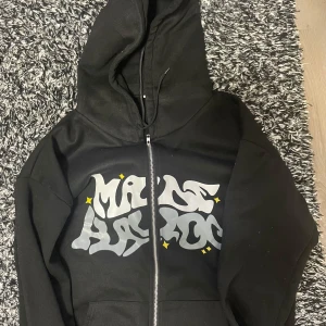 Svart made havoc hoodie - Säljer en svart hoodie med coolt tryck i grått och små gula detaljer. Den har en dragkedja framtill och en stor huva. Perfekt för en avslappnad stil.