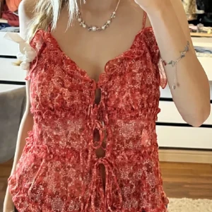 Röd blommig topp med volangdetaljer - Snygg röd blommig topp med volangdetaljer och knytning framtill. Toppen är ärmlös och har en luftig design, perfekt för varma dagar.