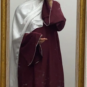 Vinröd abaya  - Elegant vinröd abaya draperad över axeln. Klänningen har dekorativa kanter och en klassisk design som ger en tidlös känsla.