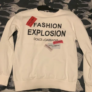 Vit sweatshirt från Dolce & Gabbana - Säljer en vit sweatshirt från Dolce & Gabbana med texten 'Fashion Explosion' tryckt på framsidan. Tröjan har långa ärmar och en klassisk rund halsringning. Perfekt för en stilren och trendig look.